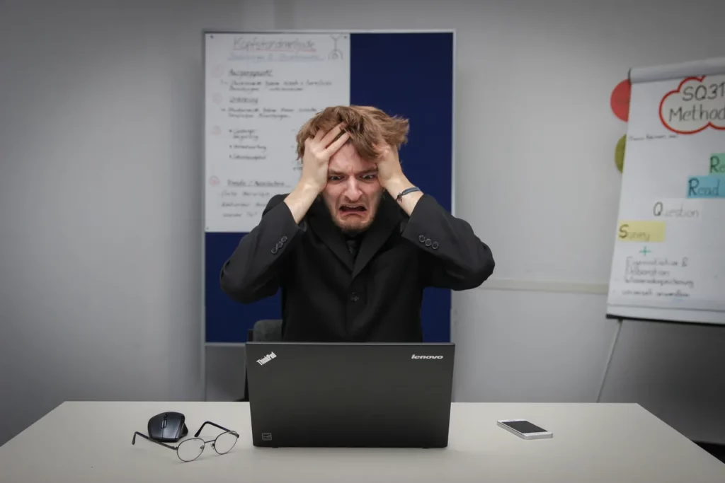 Man holding head, appearing frustrated, sits at desk with laptop.