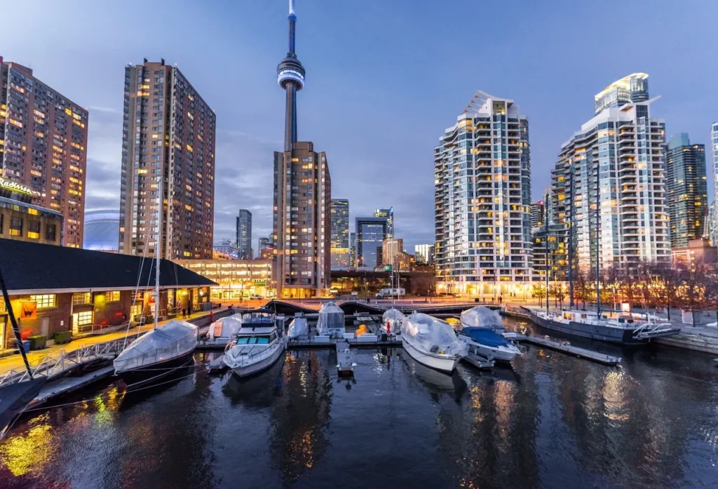 Evening cityscape with tall buildings, boats at a marina.