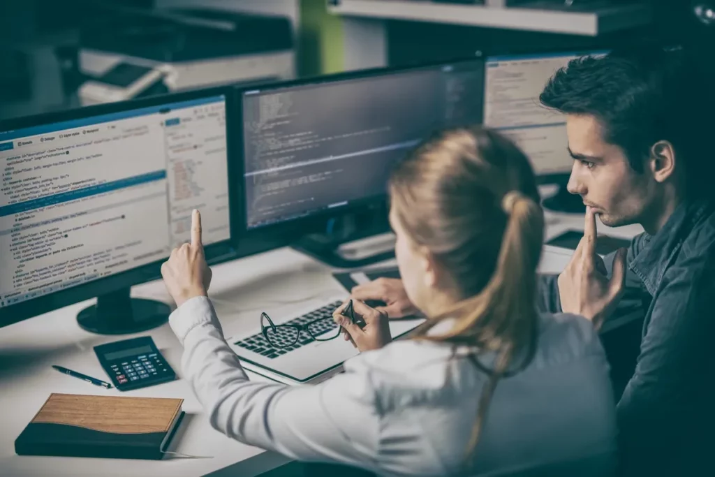 Two people discuss code on multiple computer screens.