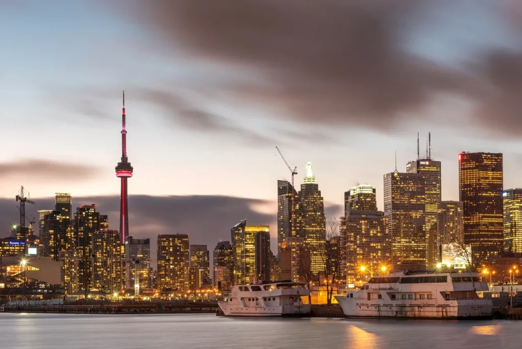 Skyscrapers glowing at dusk with boats on calm waterfront.