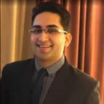 Smiling person in a suit, standing indoors near curtains.